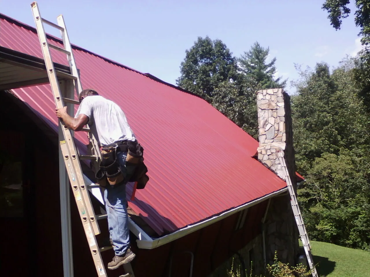 Roofer on a ladder beside a red metal roof and stone chimney — Soffit & Fascia Repair by Roofing Brothers in Laclede